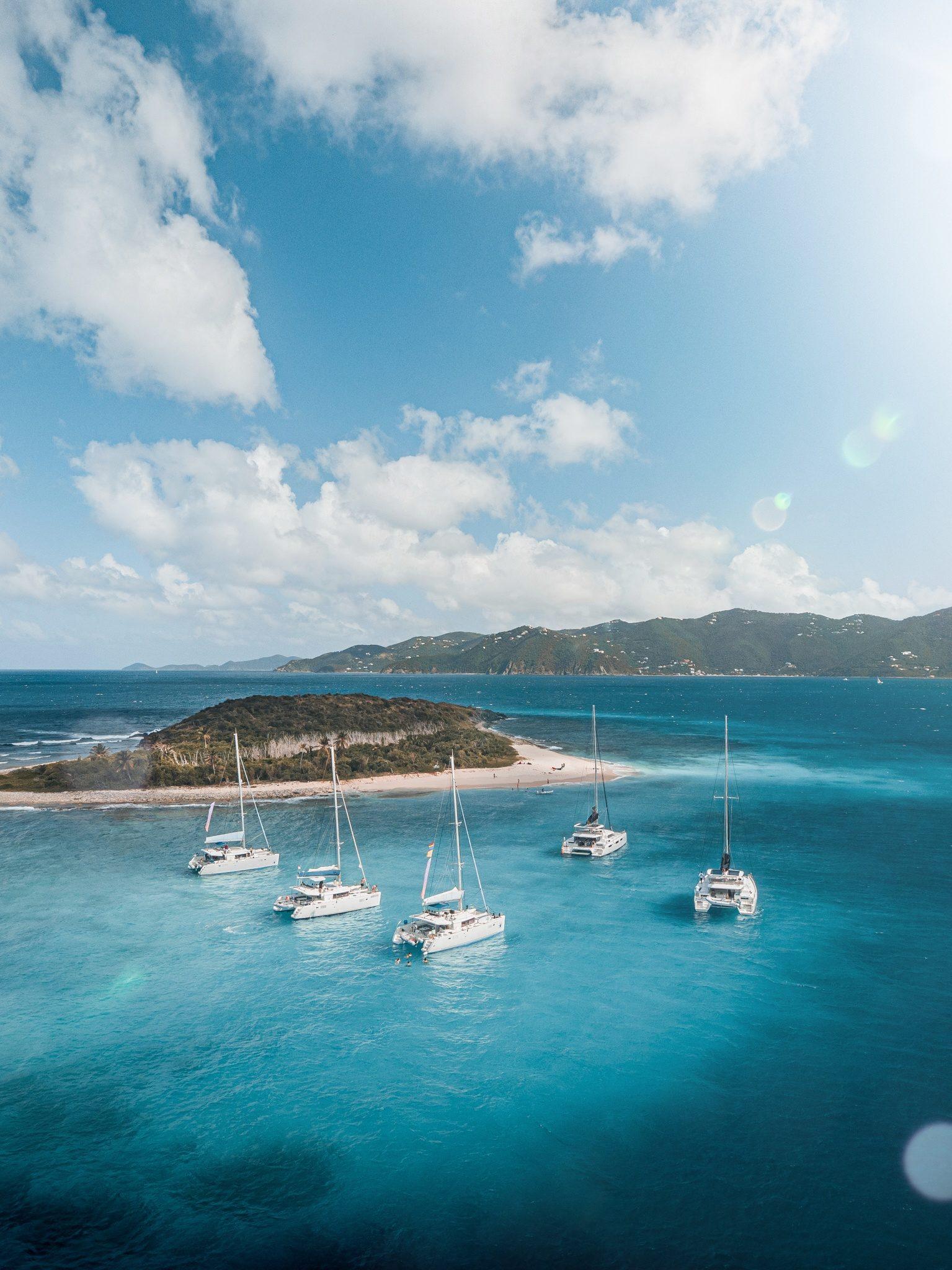 A candid, high-energy photograph of young adults jumping off a dock into clear blue ocean waters with sailing yachts anchored in the background.