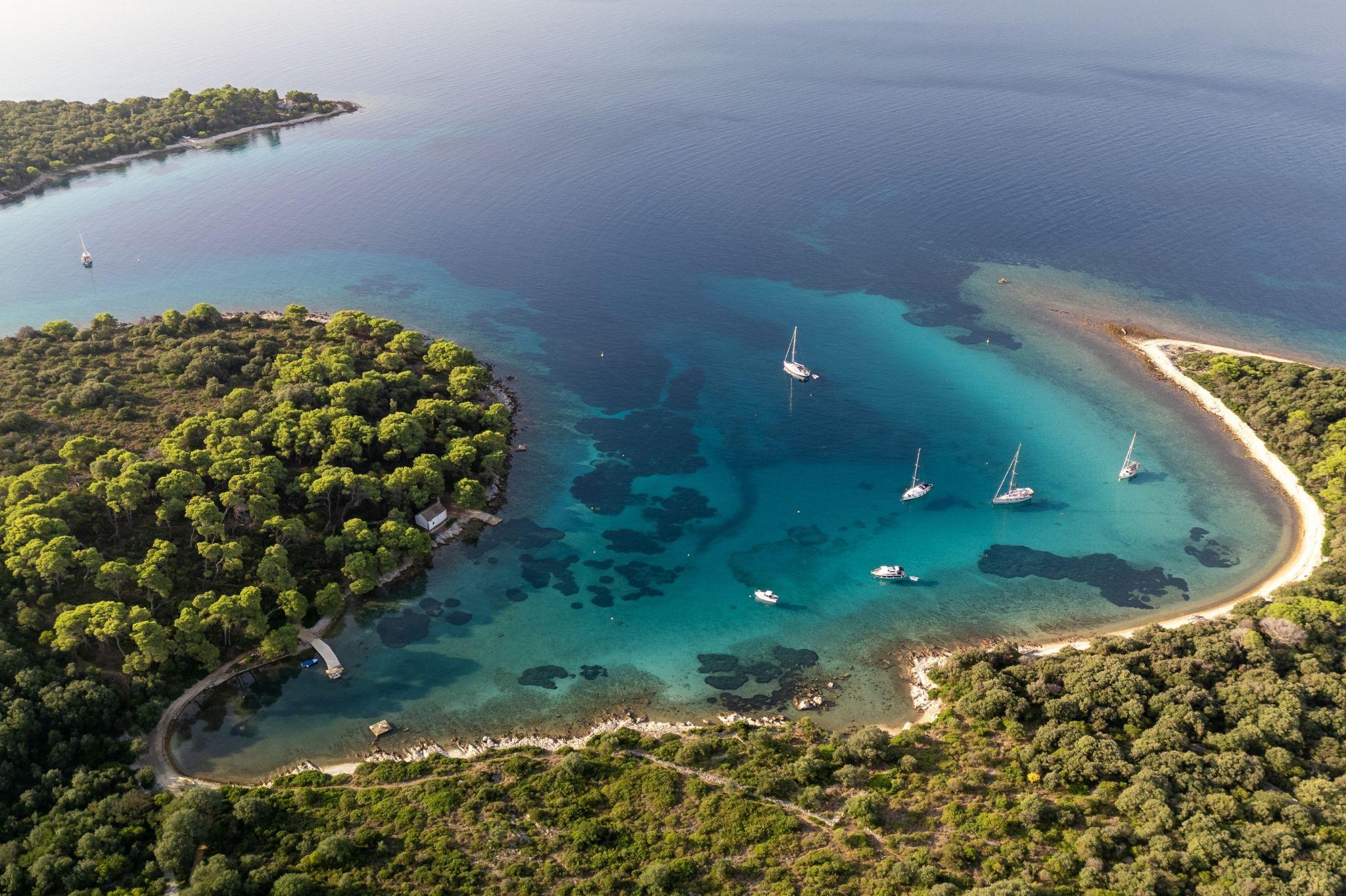 Aerial view of a scenic bay with clear blue water, sailboats, and lush green coastline.